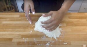 Person kneading dough on a wooden surface, preparing homemade bread in a kitchen setting.